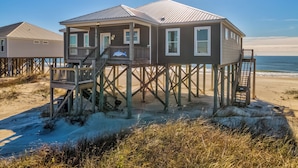 A wooden beach house with a balcony, located on stilts over the sand, with the ocean in the background.