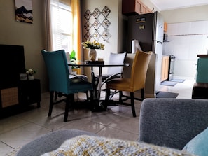 A dining area with a table and chairs, a vase with flowers, and a kitchen in the background.