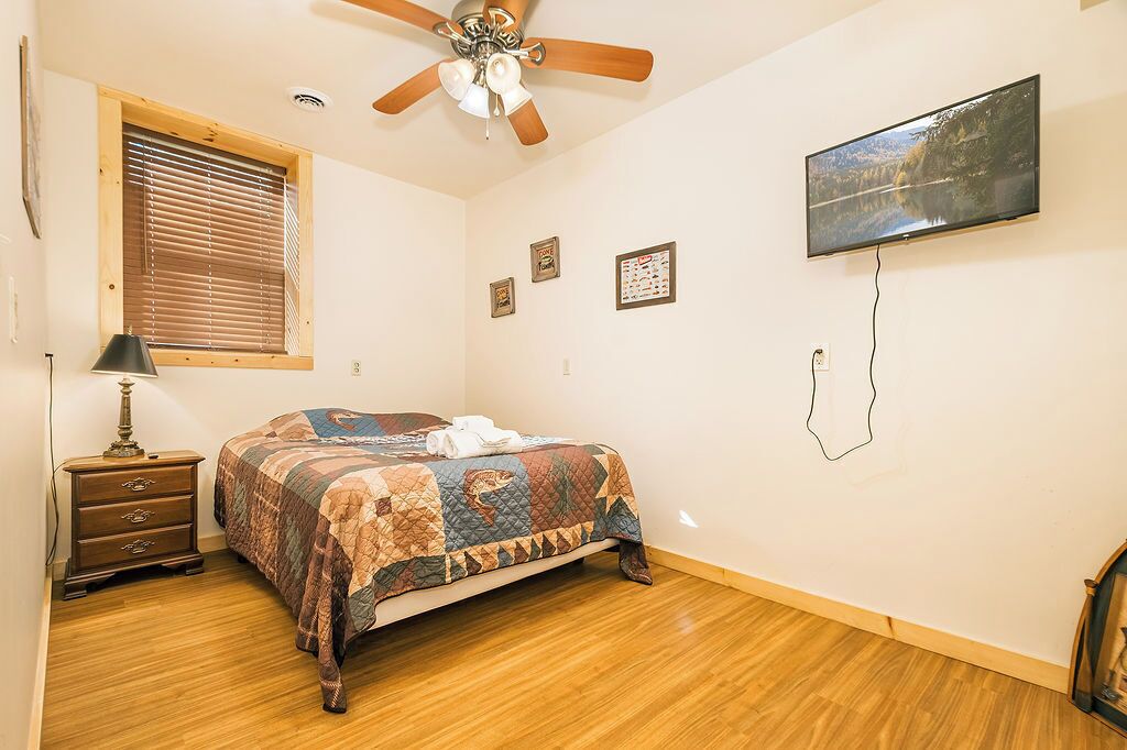 A bedroom with a bed, wooden floor, ceiling fan, window with blinds, wall-mounted TV, and a dresser.