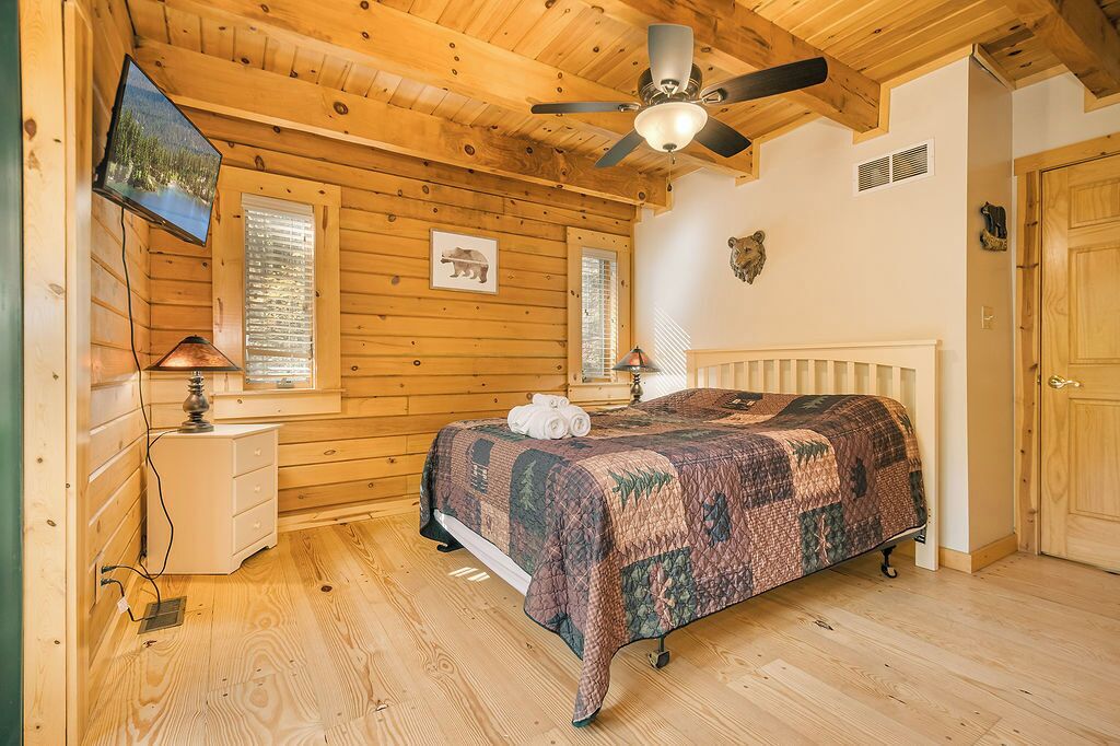 A wooden cabin bedroom with a bed, a ceiling fan, a television, and a window with blinds.