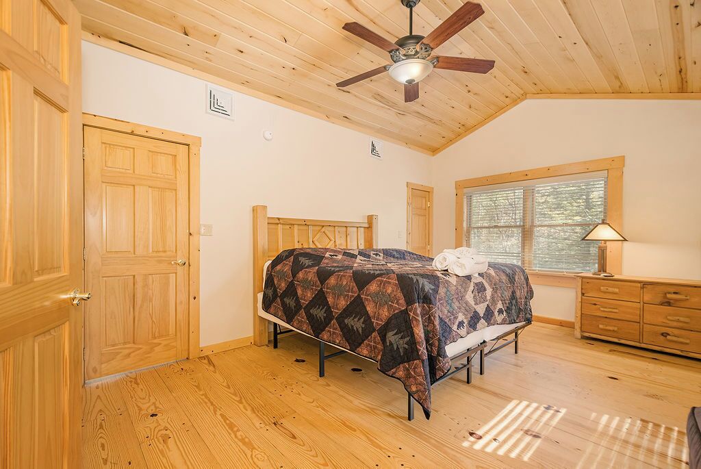Un dormitorio con techo de madera, una cama con colcha, un aparador de madera y una ventana con vista a árboles.