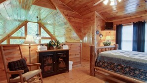 Ein gemütliches Cabin-Zimmer mit schrägem Dach, einem Bett, einem Schaukelstuhl, einer Kommode und einem Fenster mit Blick auf Bäume.