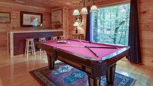 Ein Billardtisch mit roter Spielbahndecke, umgeben von hölzernen Barhocker, in einem Raum mit Holzwänden und einem großen Fenster mit Blick auf Bäume.