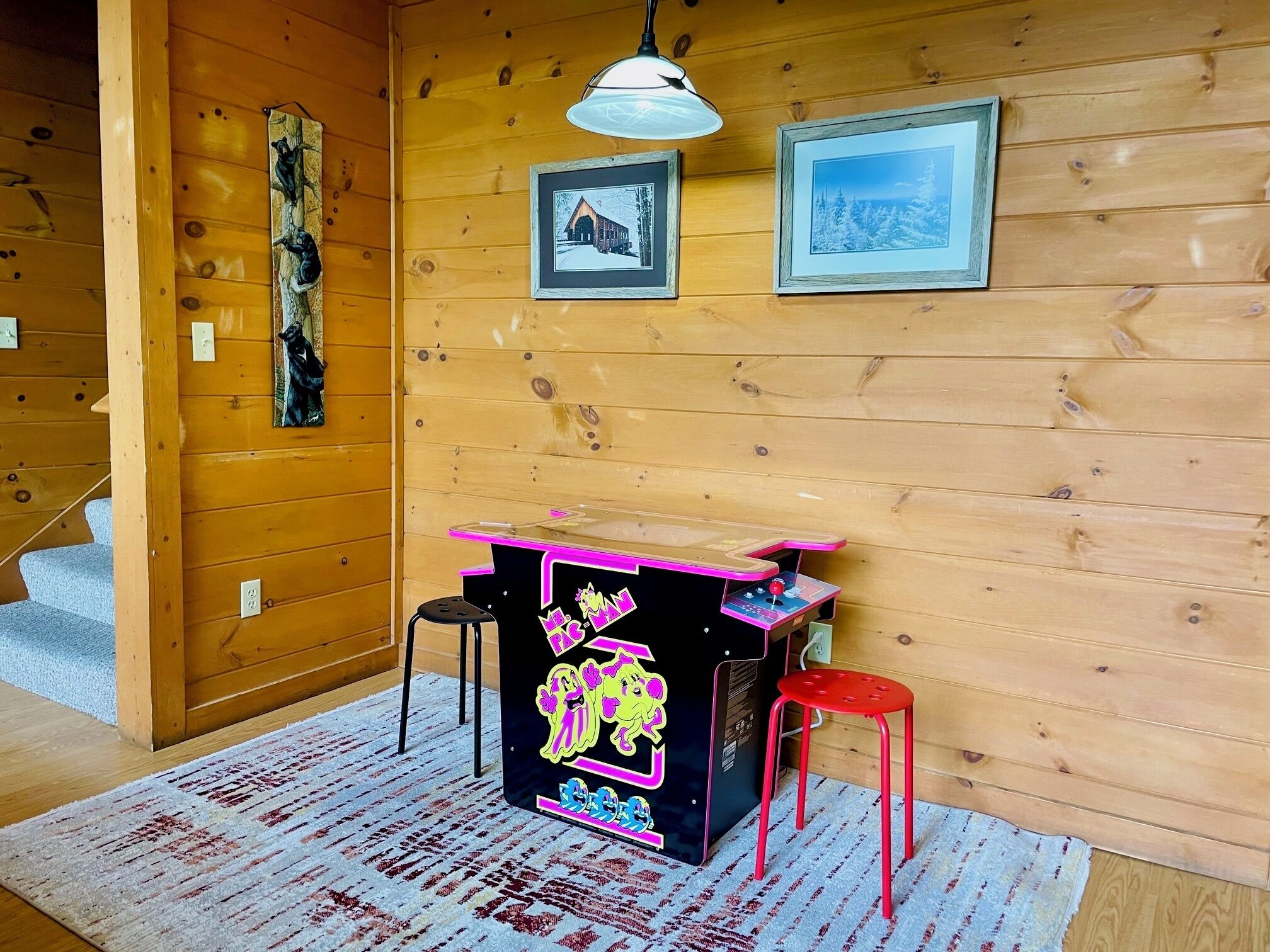 L’intérieur d’un chalet en bois, avec une borne d’arcade au néon, deux tabourets et des tableaux encadrés accrochés au mur.
