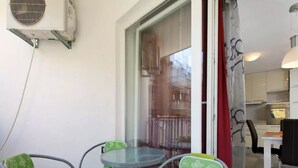 Un balcón con mesa y sillas de vidrio, vista a un edificio y una zona de cocina con armarios blancos.