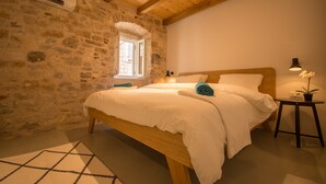 A bedroom with a stone wall, wooden ceiling beams, a bed with white bedding, a small bedside table with a lamp, and a window with a view. - Casa Sepia in Trogir old town for up to 6 people (Trogir)