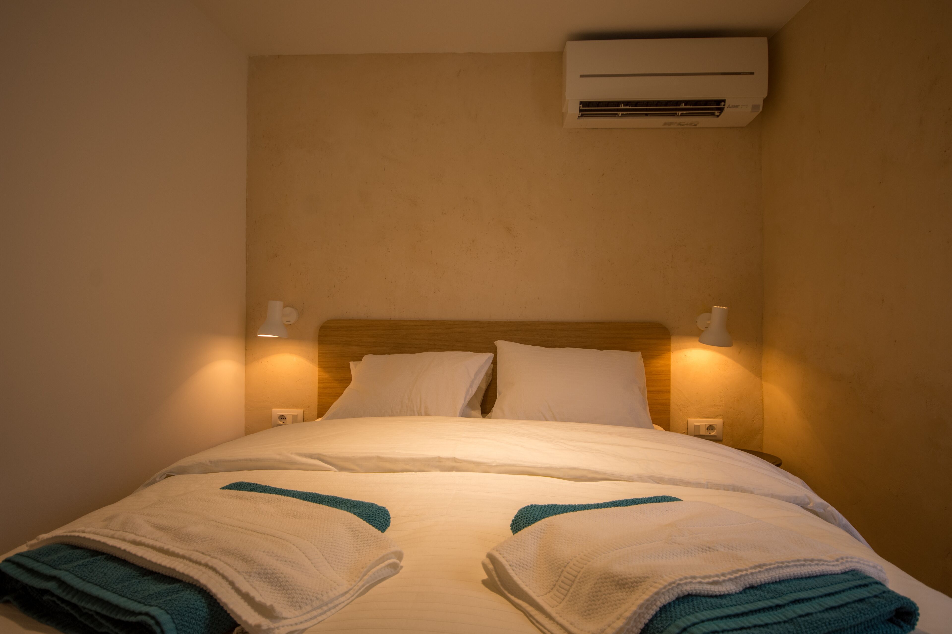 Een hotelkamer met een bed, witte lakens, een houten hoofdeinde, twee bedlampen en een airconditioner aan de muur.