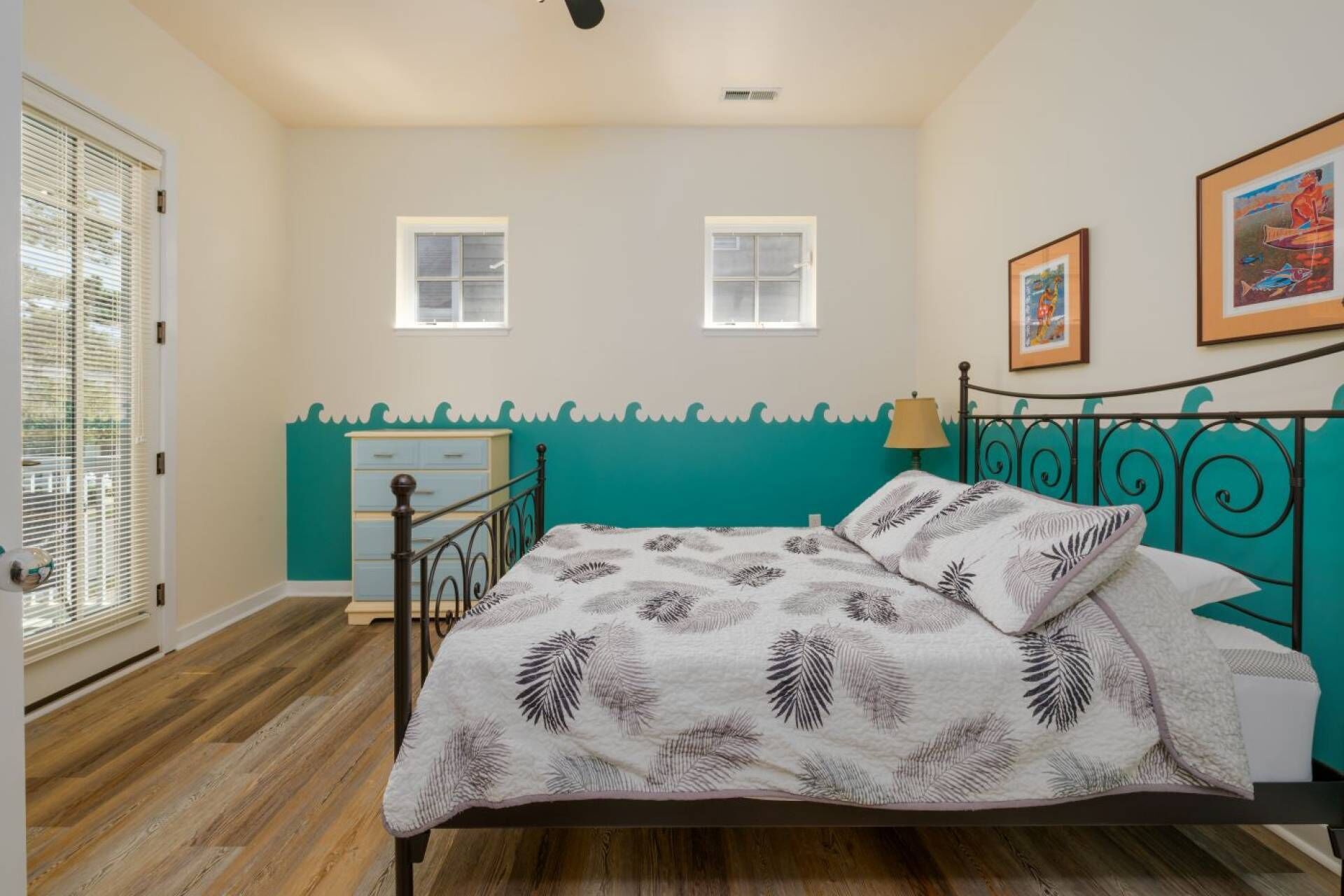A bedroom with a bed, two windows, a dresser, and a wall with a painted design.