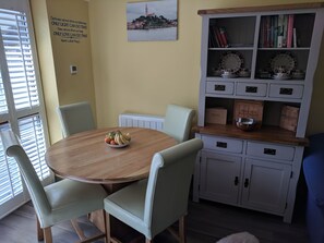 A dining area with a round wooden table, light green upholstered chairs, and a white cabinet with shelves containing books and decorative items.