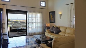Ein Wohnzimmer mit beigefarbenem Sofa, einem Couchtisch und Blick auf den Strand durch eine Glastür.