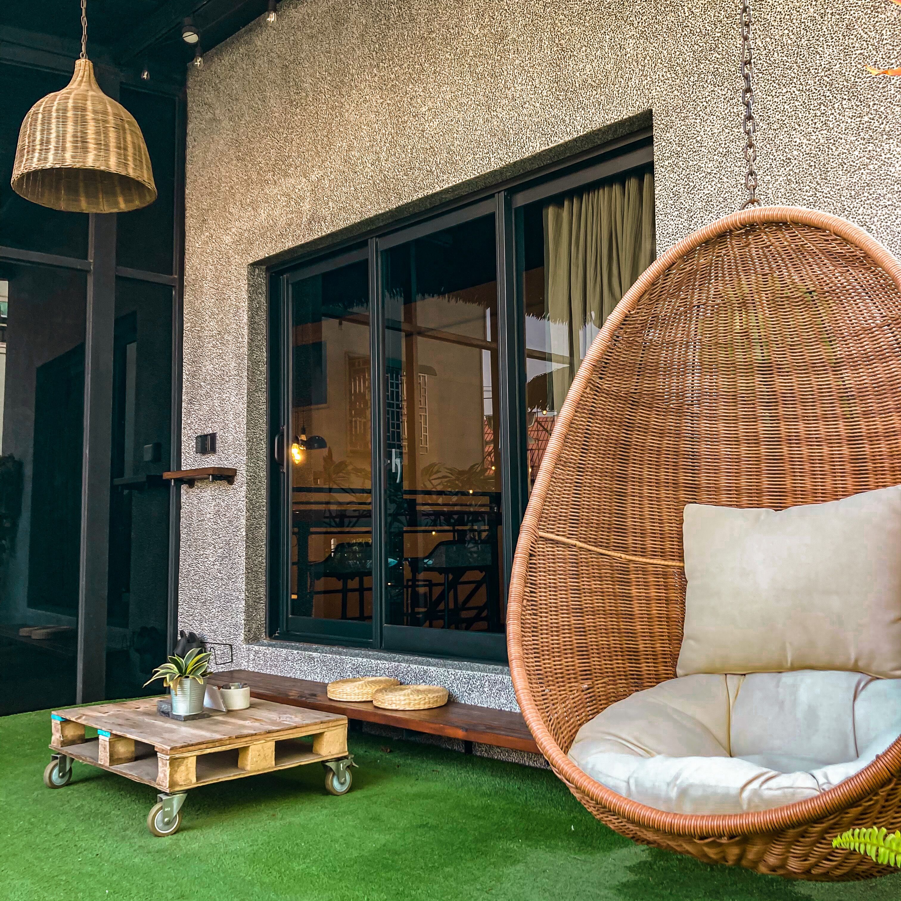 A wicker chair with a cushion, a wooden cart with a potted plant, and a large window with curtains.