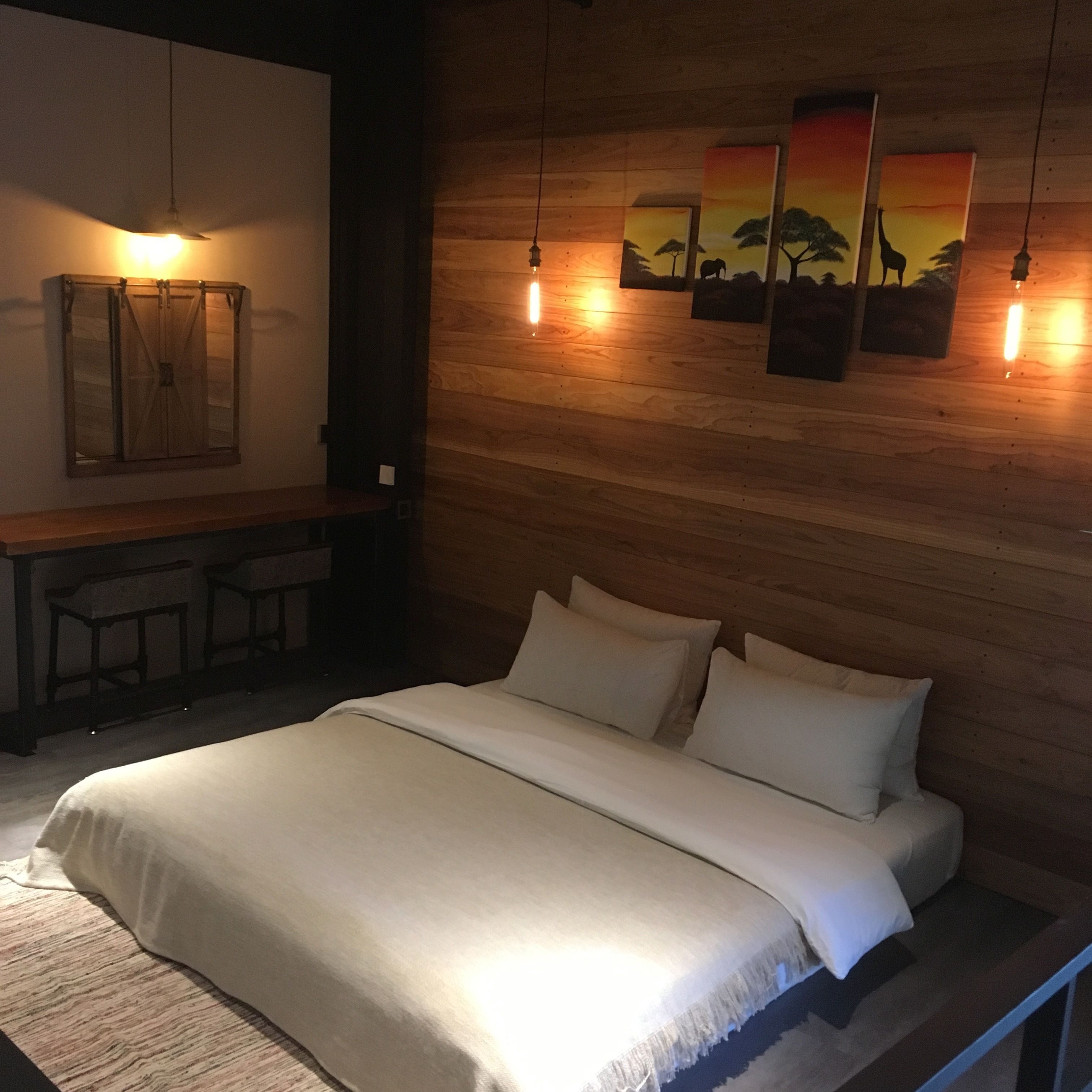Une chambre à coucher avec une tête de lit en bois, un lit recouvert de draps blancs, un bureau avec deux chaises et des œuvres d’art accrochées au mur.