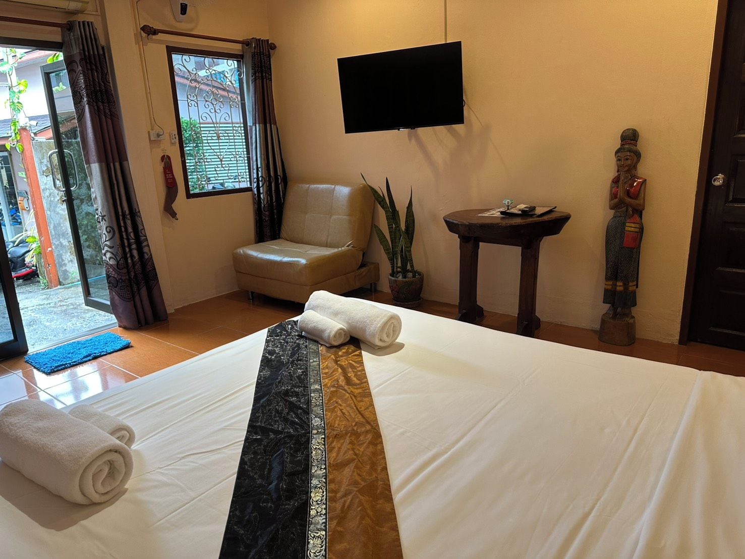 Habitación de hotel con cama, una silla, televisor, mesita, planta en maceta y una estatua.