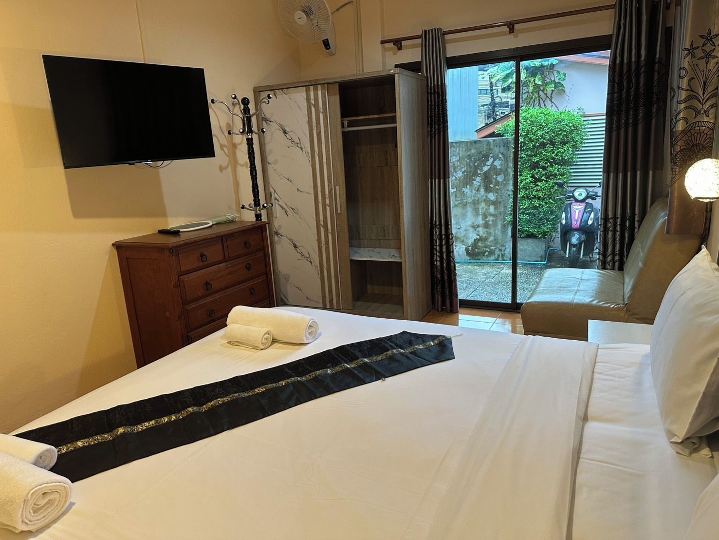Habitación de hotel con cama, televisor montado en la pared, cómoda de madera, puerta corrediza de vidrio que da a un área exterior y ventilador de techo.