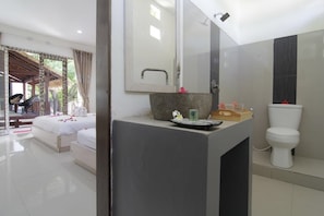 A modern bathroom with a large mirror, a stone sink, and a toilet. There is a bed with white linens and a small table with toiletries. - Gili Ilalang Village (Gili Trawangan)