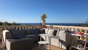 A sunlit outdoor seating area with a sectional sofa, two small round tables, and a cart with drinks and snacks. A palm tree and a clear view of the sea are visible in the background. - Lirios Boutique Luxury Seaview Villa ⭐︎⭐︎⭐︎⭐︎⭐︎ (Benitachell)