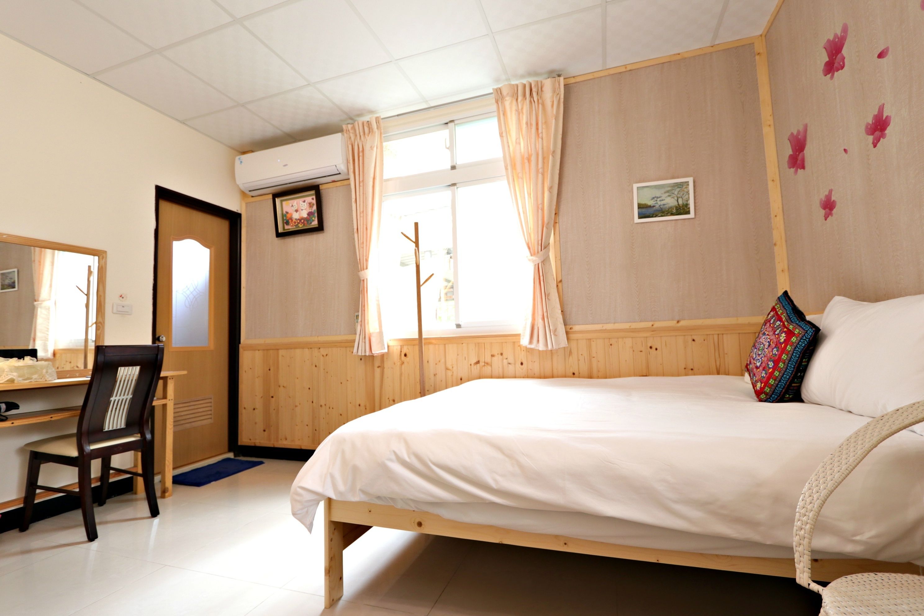 Een slaapkamer met een houten bed, een bureau met stoel, een raam met gordijnen en een luchtverkoeler aan de muur.