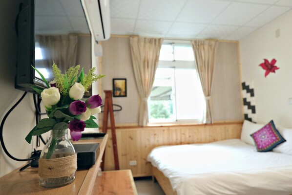 A bedroom with a bed, a wooden desk, a small table with a vase of flowers, a TV mounted on the wall, and a window with curtains.