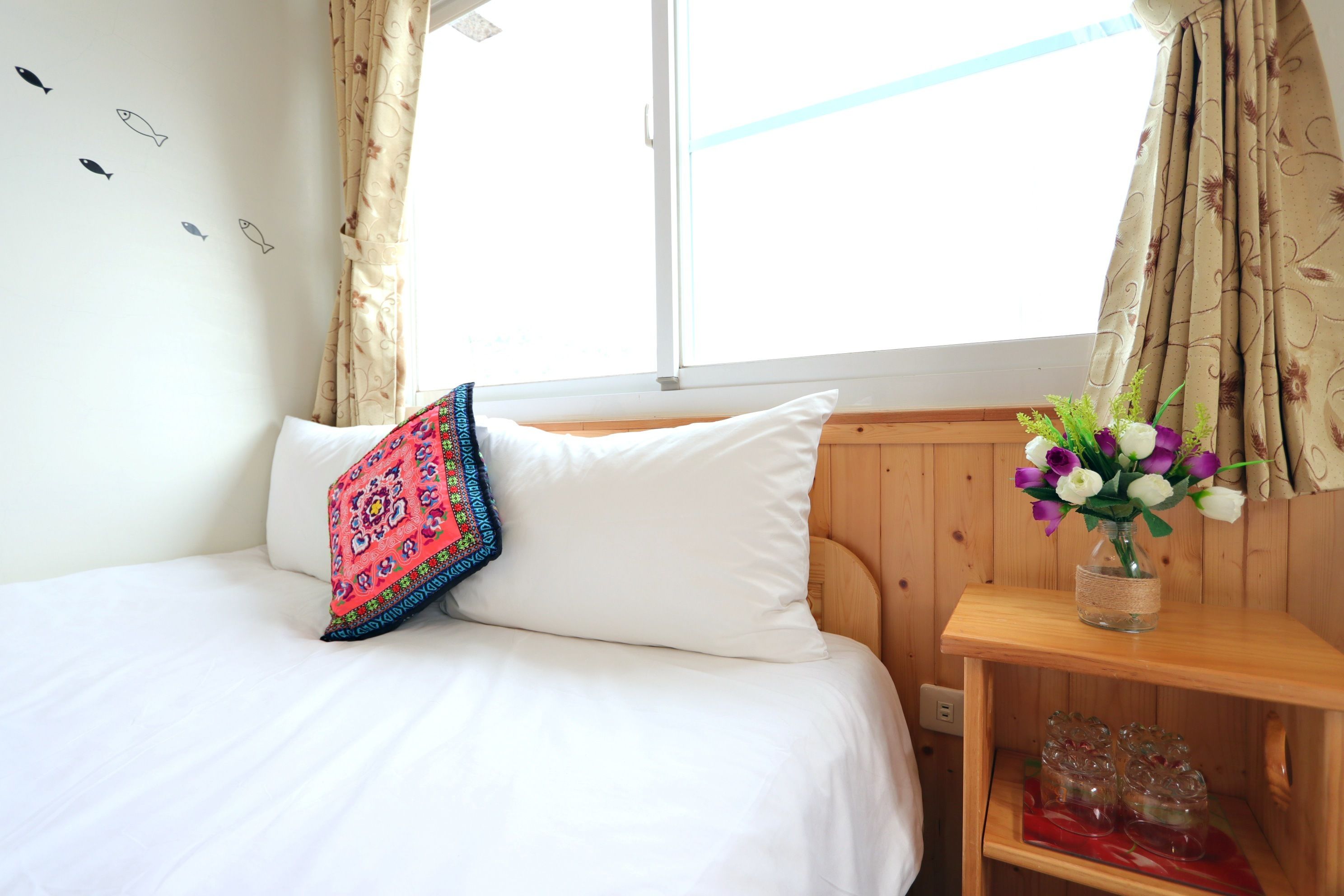 Un dormitorio con una cama, un cojín colorido, un florero con flores y una mesita de noche de madera.