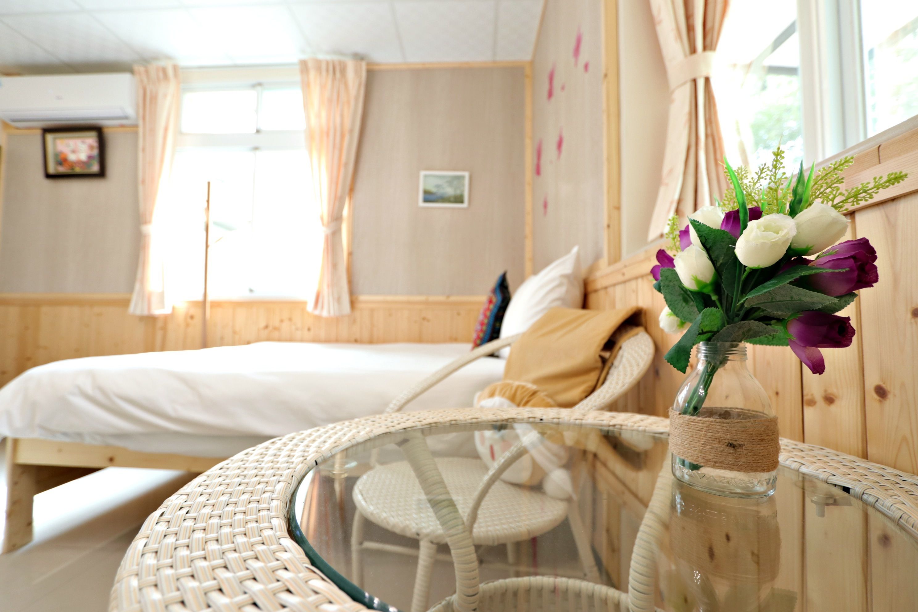 Una habitación con una cama, una silla de mimbre y una mesa de vidrio con un florero.