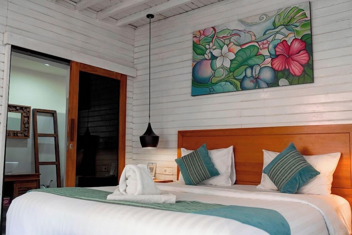 A hotel room with a wooden headboard, a bed with white linens, a painting of tropical flowers, and a wooden door.