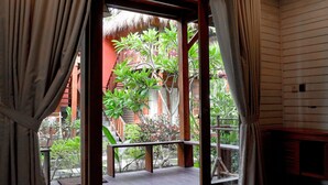 A room with a wooden floor, a window with curtains, and a view of a red building and greenery outside.