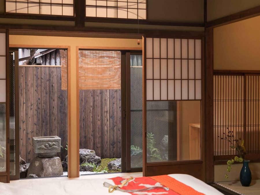 Ett traditionellt japanskt rum med tatamimattor på golvet, en skjutbar shoji-skärmdörr, en stenlykta och en säng med rött och vitt sänglinne.