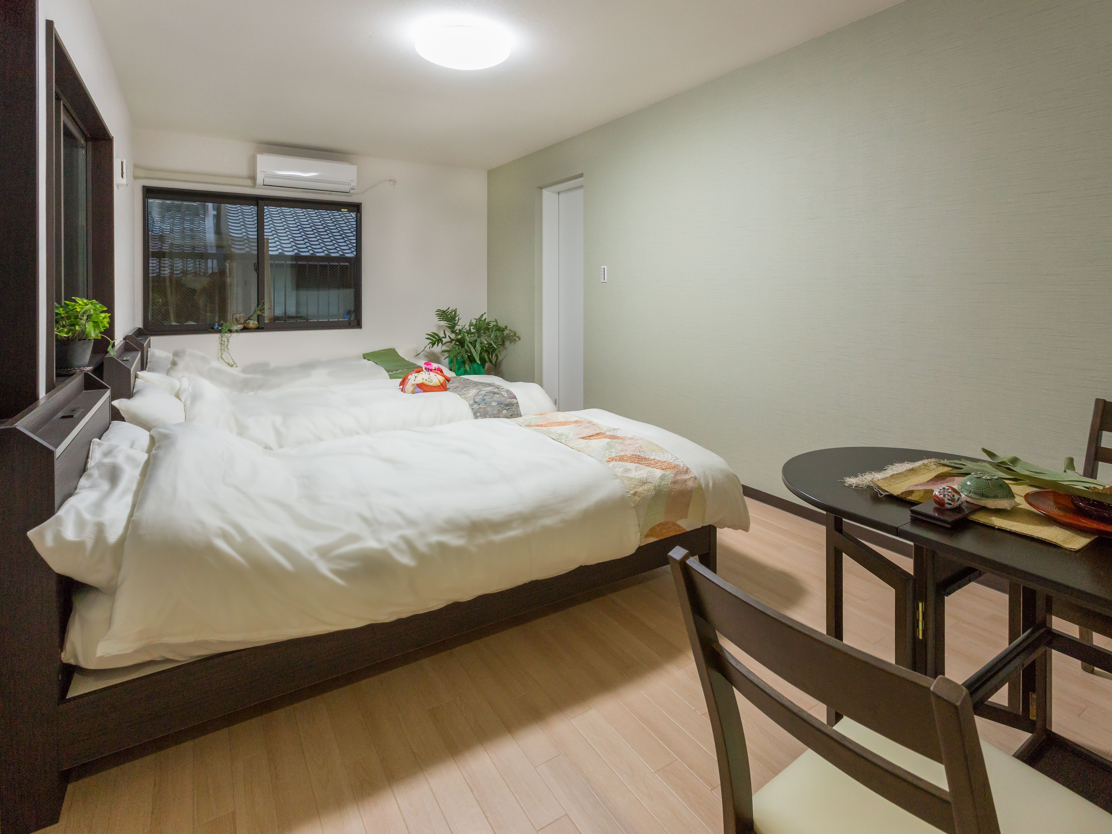 A neatly made bed with a white comforter and pillows, a small table with a placemat and a teapot, and a chair.