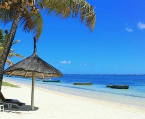 A beach with a thatched umbrella, palm trees, and boats in the water. - Private Bungalow - Beach Front (Flic en Flac)