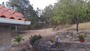 Un paysage rural avec un chemin de terre, un mur de pierres agrémenté de plantes en pot et un bâtiment surmonté d’un toit de tuiles rouges.