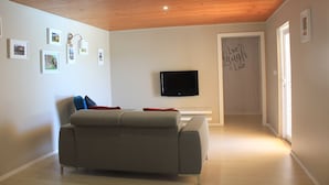 A living room with a grey sofa, a flat-screen TV mounted on the wall, and a door with a sign that says "We Laugh Love".