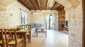 Une pièce aux murs de pierre et au plafond en bois, comprenant un coin repas avec des chaises et une table, un canapé et un téléviseur posé sur une étagère en bois.