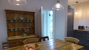 A wooden dining table with a bowl of fruit, wooden chairs, a wooden shelf with cups, and a kitchen area with cabinets and a toaster. - A delightful country house set in the heart of Lakeland (Keswick)