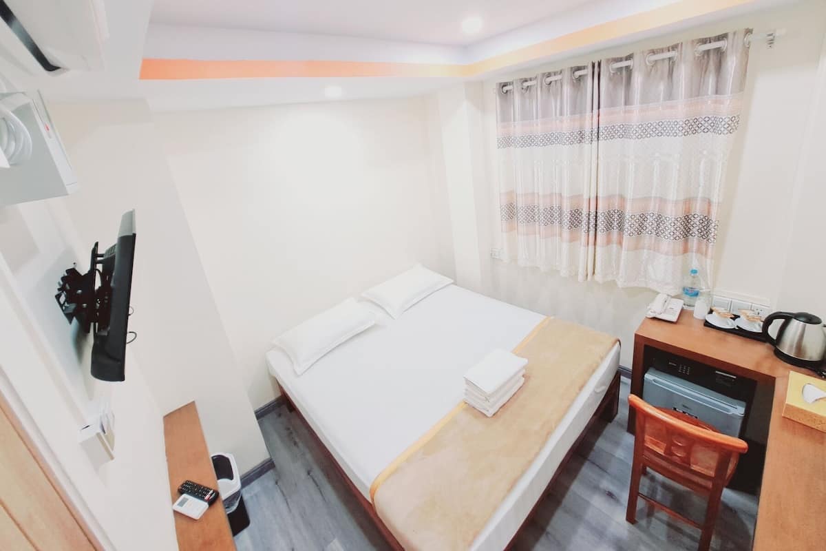 A hotel room with a bed, a desk with a chair, a small table, a kettle, and a window with curtains.