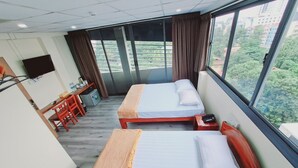 A hotel room with two beds, a desk with a chair, a TV mounted on the wall, and a large window with a view of buildings and greenery. - 121 Hotel Yangon (Yangon)