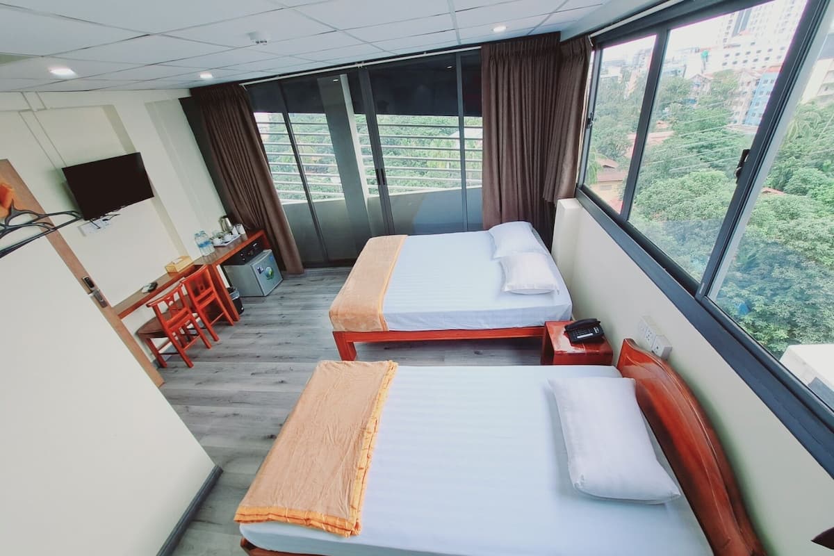 A hotel room with two beds, a desk with a chair, a TV mounted on the wall, and a large window with a view of buildings and greenery.