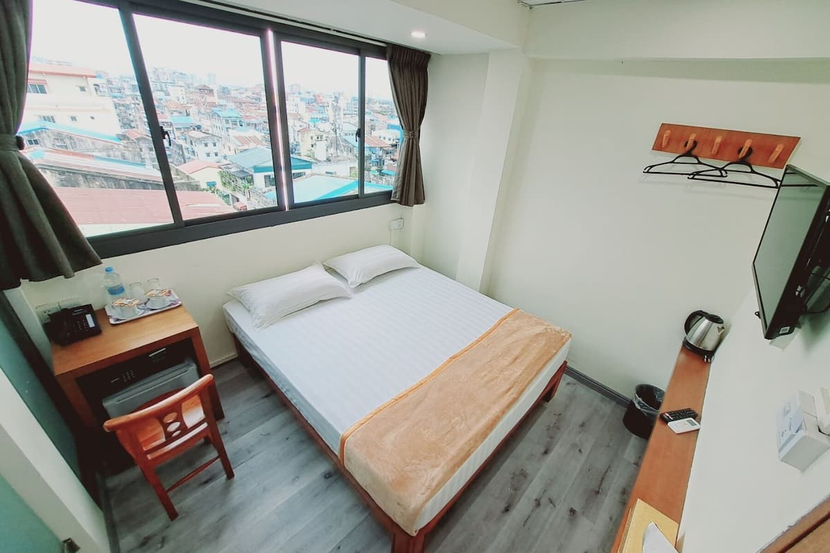 A hotel room with a bed, a desk with a chair, a TV, and a view of the city through large windows.