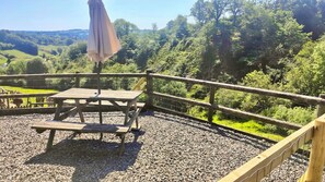 A wooden deck with a picnic table and an umbrella, surrounded by a wooden fence and gravel, overlooking a forested landscape.