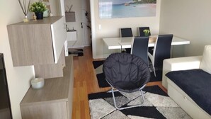 A modern living room with a sofa, a black chair, a dining table with chairs, a kitchen area, and a painting of a beach scene.