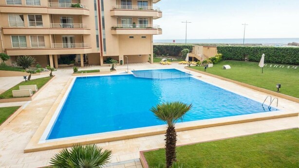 A swimming pool surrounded by a well-maintained lawn, a multi-story building with balconies, and a clear view of the sea.