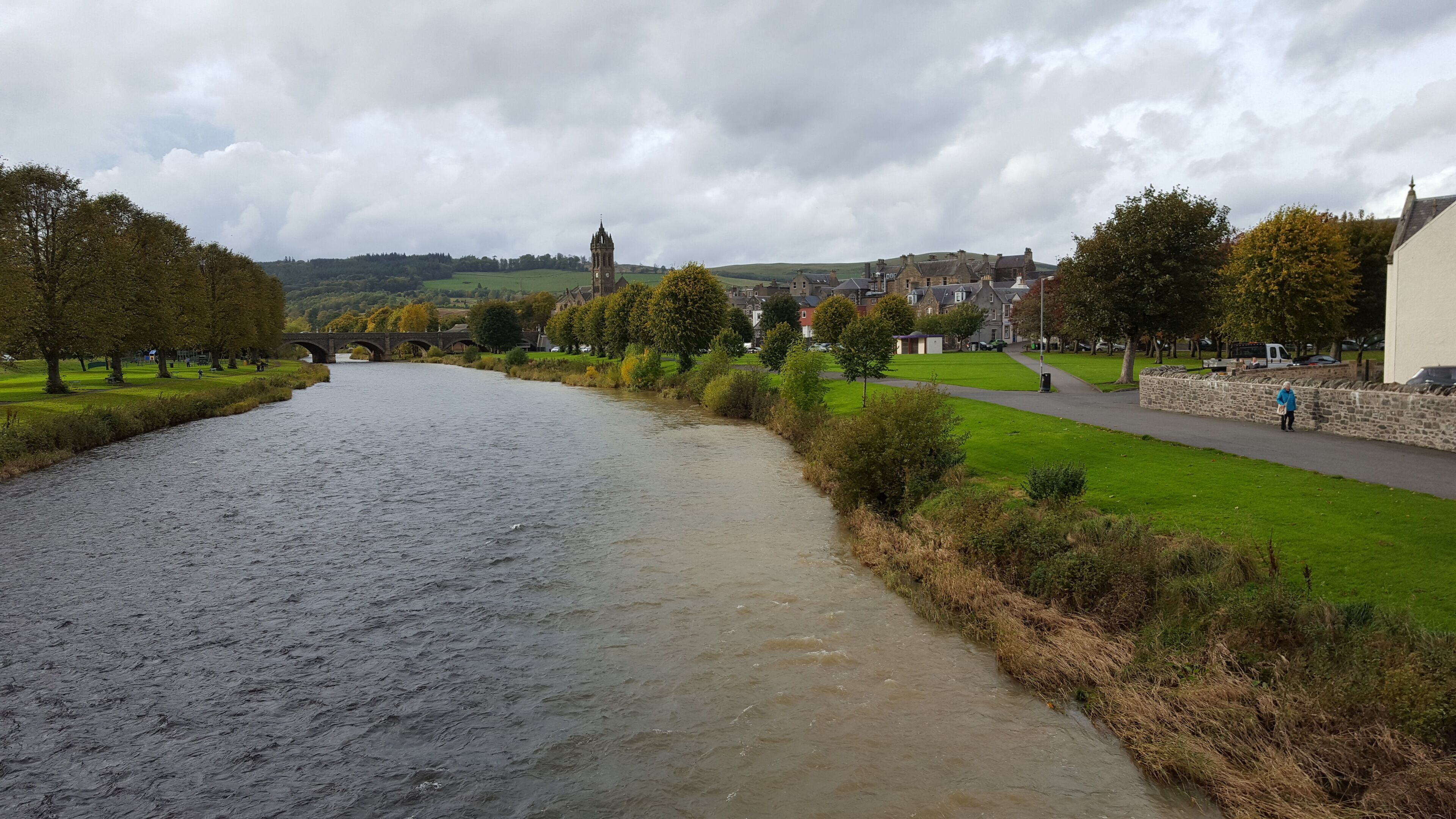 Ein Fluss mit einer Steinbrücke, einem grünen Pfad und Gebäuden im Hintergrund.