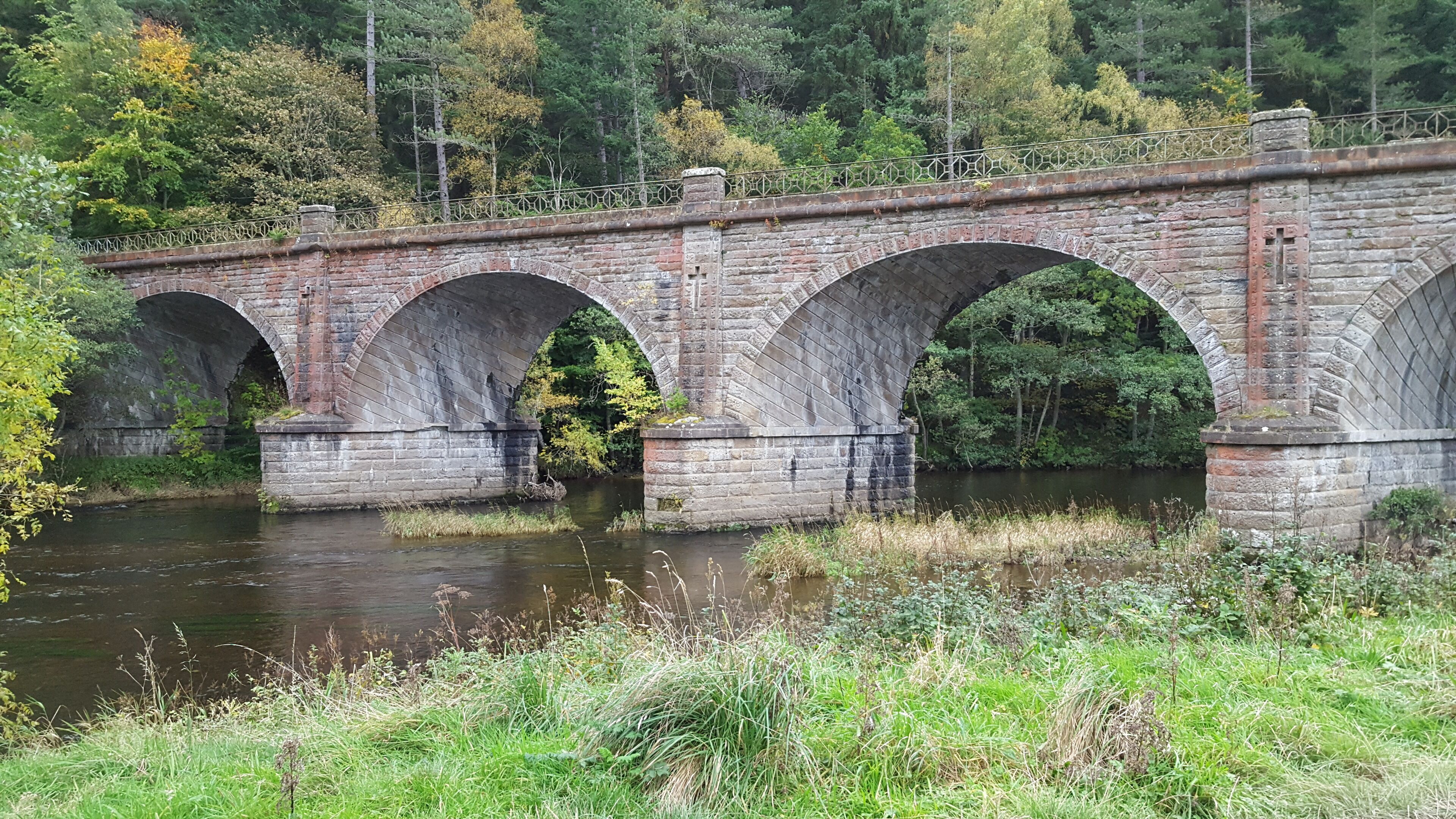 Een stenen brug met drie bogen over een rivier, omgeven door groen en bomen.