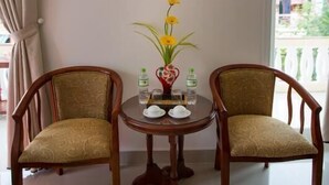 Ein Zimmer mit zwei Holzstühlen, einem kleinen runden Tisch, einer Vase mit gelben Blumen und einem Hausbild an der Wand.