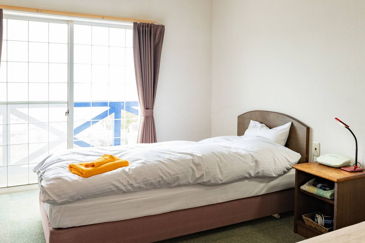 A hotel room with a bed, a bedside table with a lamp, and a view of a blue structure outside the window.