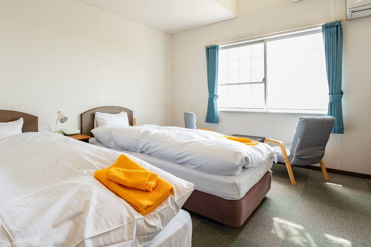 A hotel room with two beds, a chair, a window with curtains, and a bedside table with a lamp.