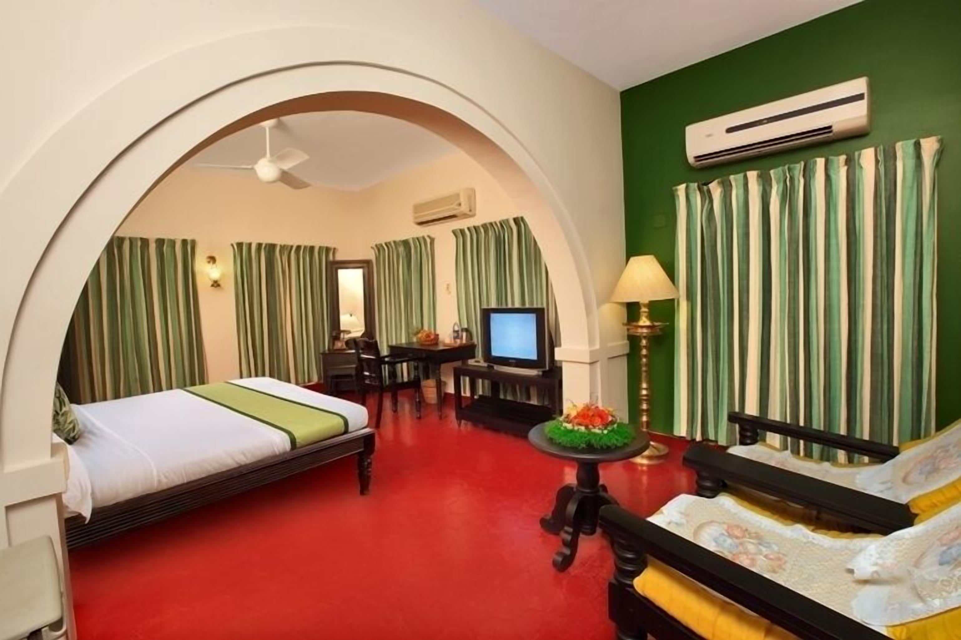 Habitación de hotel con alfombra roja, cama, escritorio con televisor, lámpara y sofá.