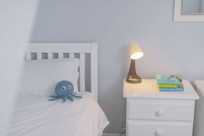 A bedroom with a white bed, a blue plush toy, a bedside lamp, and a stack of books on a nightstand.