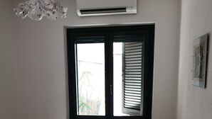 A bedroom with two single beds, a radiator, a window with blinds, a small table with a lamp, and an air conditioner. - Holiday house in residence near the sea (Borgo Hermada 29)