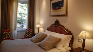 Ett hotellrum med en stor säng, två sänglampor, ett fönster med gardiner, en inramad tavla på väggen och utsikt över omgivningarna.
