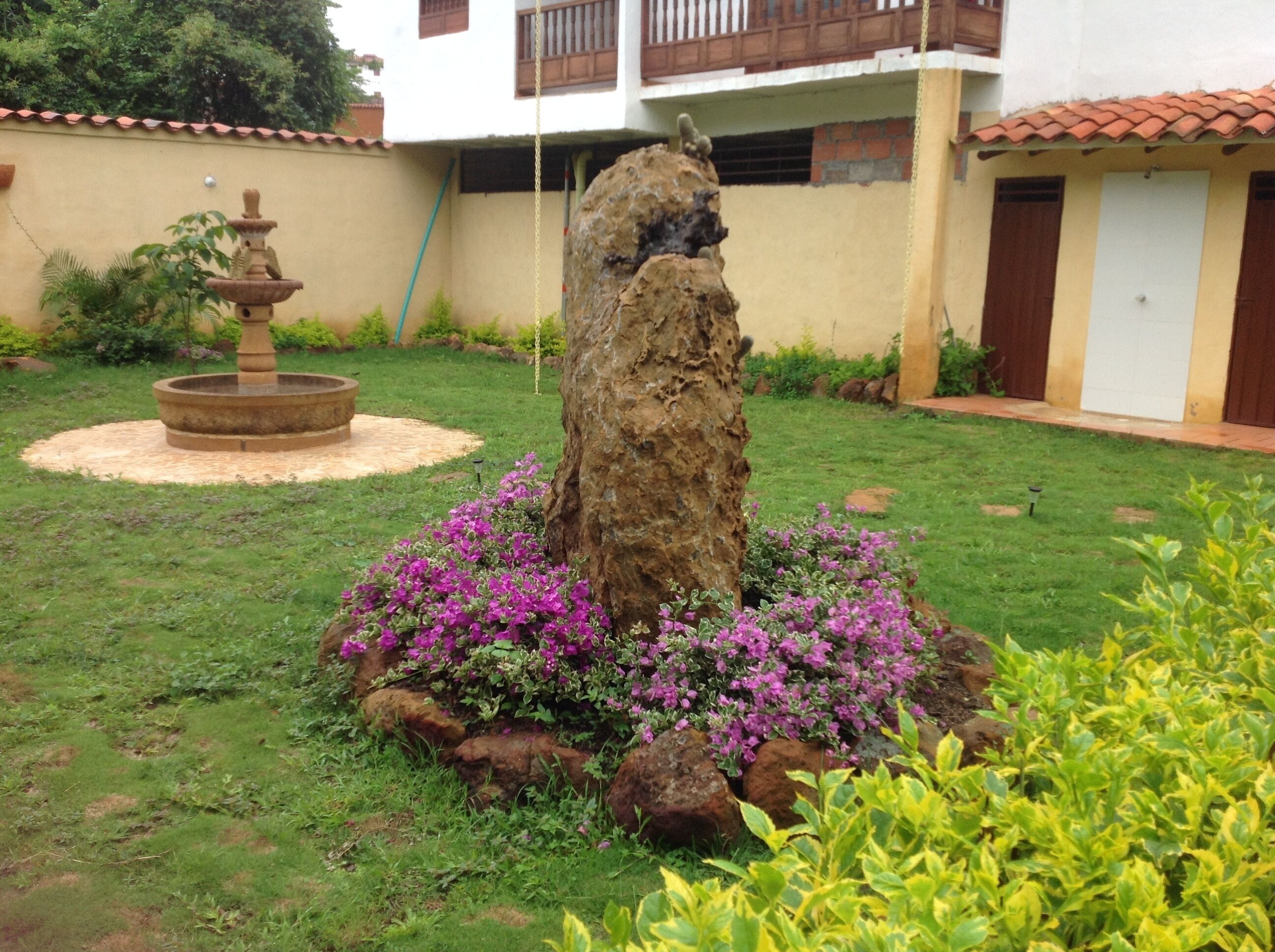 Un jardín con una formación rocosa central, una fuente y plantas florecientes.